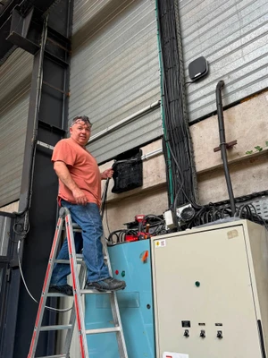 Portrait d'un technicien ou responsable d'entreprise de sécurité électronique, âgé d'environ 45-55 ans, portant des vêtements professionnels (polo ou chemise avec logo FD Sécurité), photographié dans un atelier ou local technique avec équipements de sécurité visibles en arrière-plan (coffrets électriques, caméras, centrales d'alarme). L'homme regarde directement l'appareil photo avec une expression professionnelle et approachable, debout avec une posture confiante, incarnant l'expertise et l'engagement du dirigeant.