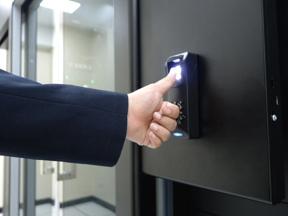 Vue rapprochée en légère plongée d'une main d'un professionnel appuyant sur le clavier rétroéclairé d'un lecteur de badge de contrôle d'accès monté en applique murale. Boîtier en aluminium brossé avec pavé numérique à touches bleues illuminées, écran LCD affichant un statut d'accès, voyant LED vert de validation, et lecteur RFID intégré. En arrière-plan flou, une porte de sécurité avec cadre métallique. L'image capture le geste professionnel d'activation d'un accès paramétré, mettant en avant la précision et le contrôle de la sécurité des zones.