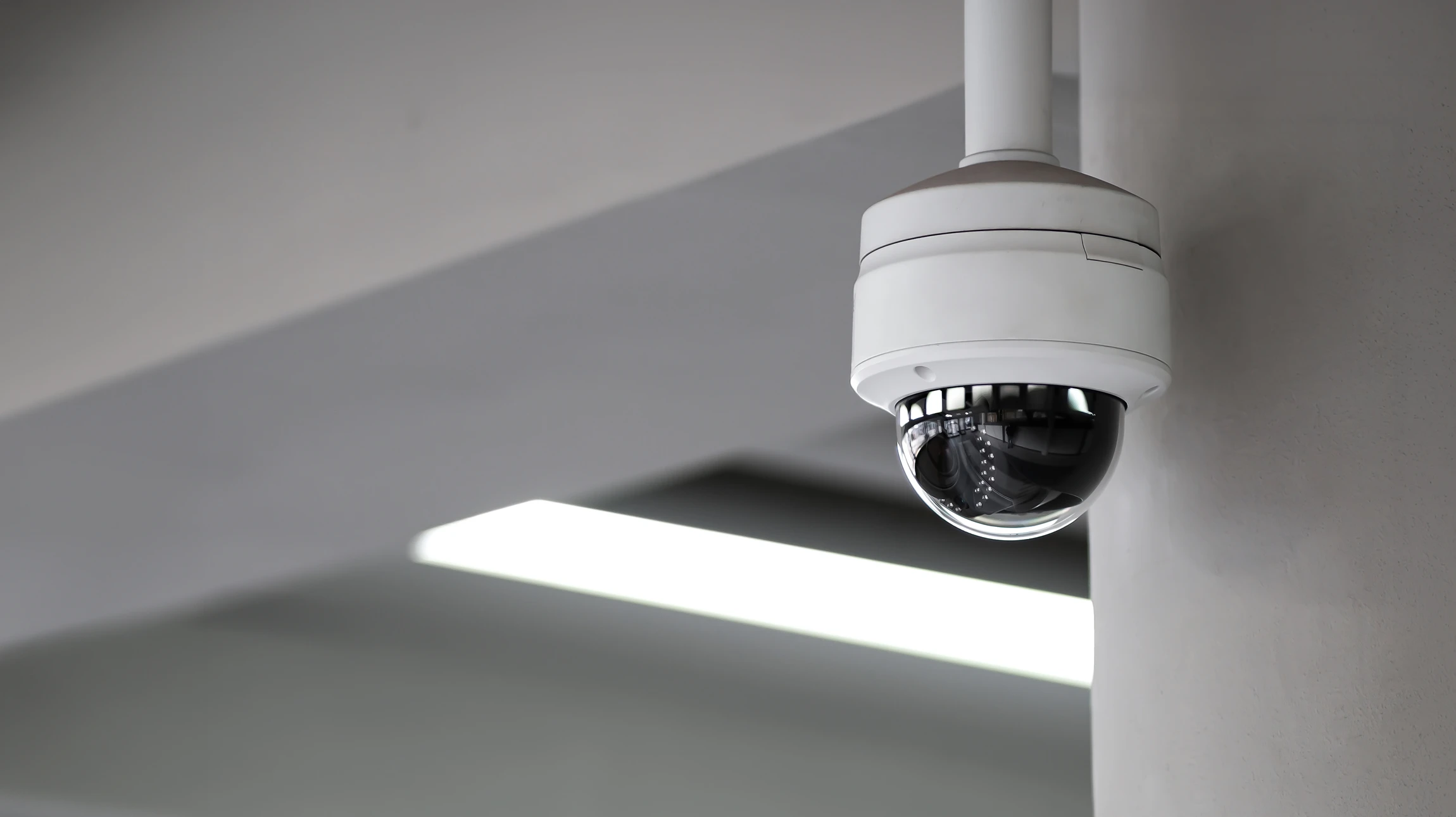 Caméra de surveillance extérieure moderne montée sur mur blanc, boîtier cylindrique blanc avec extrémité noire, arrière-plan flou avec effet bokeh lumineux suggérant un environnement urbain nocturne