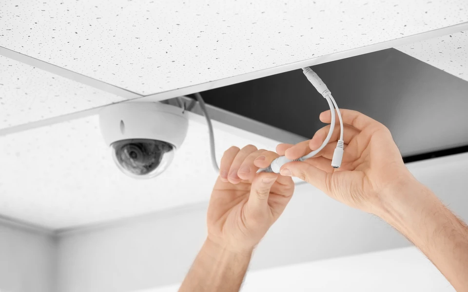 Technicien en combinaison de travail grise tenant plusieurs caméras de vidéosurveillance blanches et outils de maintenance dans un atelier intérieur