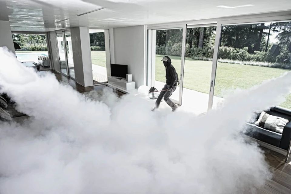 Photo réaliste en intérieur professionnel montrant un générateur de brouillard monté discrètement en plafond ou en hauteur dans un local commercial ou entrepôt. Le boîtier, compact et discret, est visible avec ses sorties de diffusion. Une légère brume opaque remplit progressivement l'espace dans l'arrière-plan de l'image, créant une visibilité très réduite qui illustre l'effet de barrière immédiate. L'éclairage professionnel crée un contraste permettant de bien percevoir le phénomène de blocage visuel. Style photo professionnelle de sécurité, sans texte ni logo apparent.